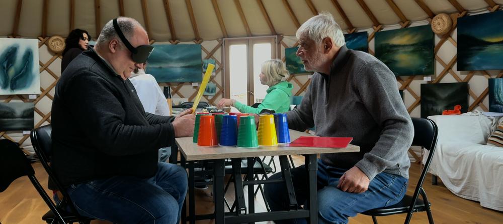Two participants at a table of coloured cups; one wears a blackout mask while the other guides the exercise.